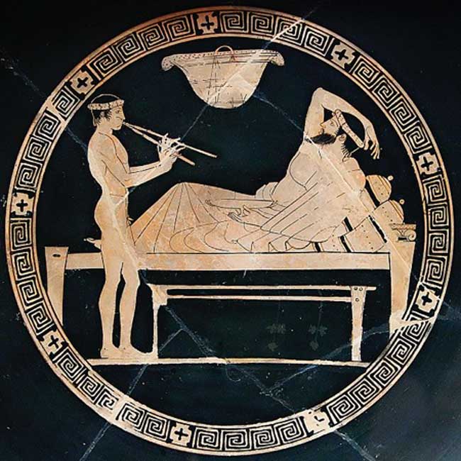 Escena de un banquete: un hombre adulto se reclina sobre un diván mientras un joven muchacho toca el aulós, instrumento de viento de la antigua Grecia. Tondo de una copa ática de figuras rojas (Dominio público).