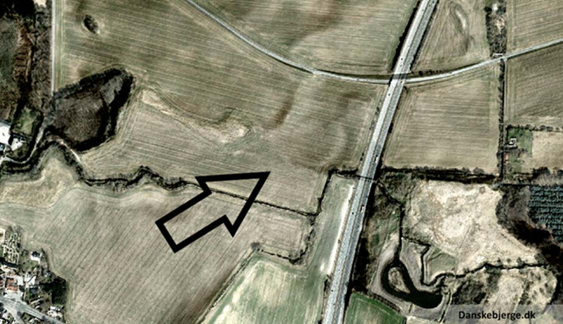Fotografía aérea de Vallø Borgring. Versión modificada de una fotografía de satélite en la que se han sombreado los relieves. La flecha señala en lugar en el que se observa una forma circular, emplazamiento de la antigua fortificación. (Danskebjerge/CC BY SA 3.0)